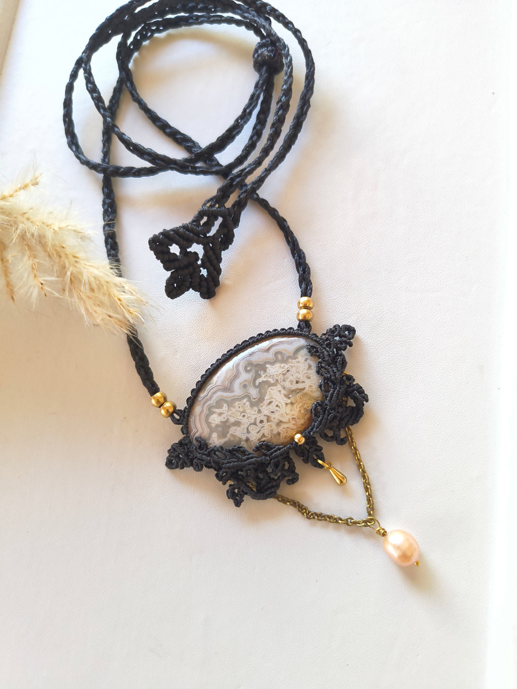 A handmade macrame pendant necklace featuring a polished crazy lace agate stone wrapped with black waxed macrame threads, gold beads, and a dangling pearl, styled on a light beige surface with dried pampas grass.