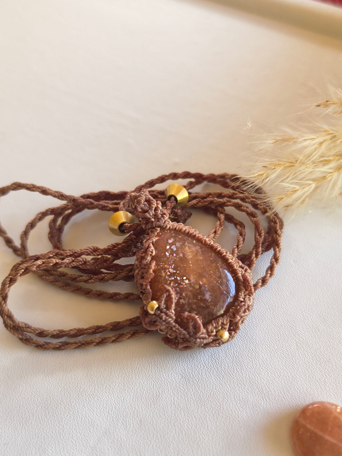A handmade macrame pendant necklace featuring a high-quality sunstone wrapped in brown braided cord, accented with small brass beads, displayed on a light beige textured surface.