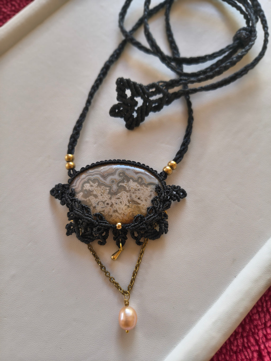 A handmade macrame pendant necklace featuring a polished agate slice, gold-tone beads, and a dangling pink freshwater pearl on a gold chain, displayed on a white tray.