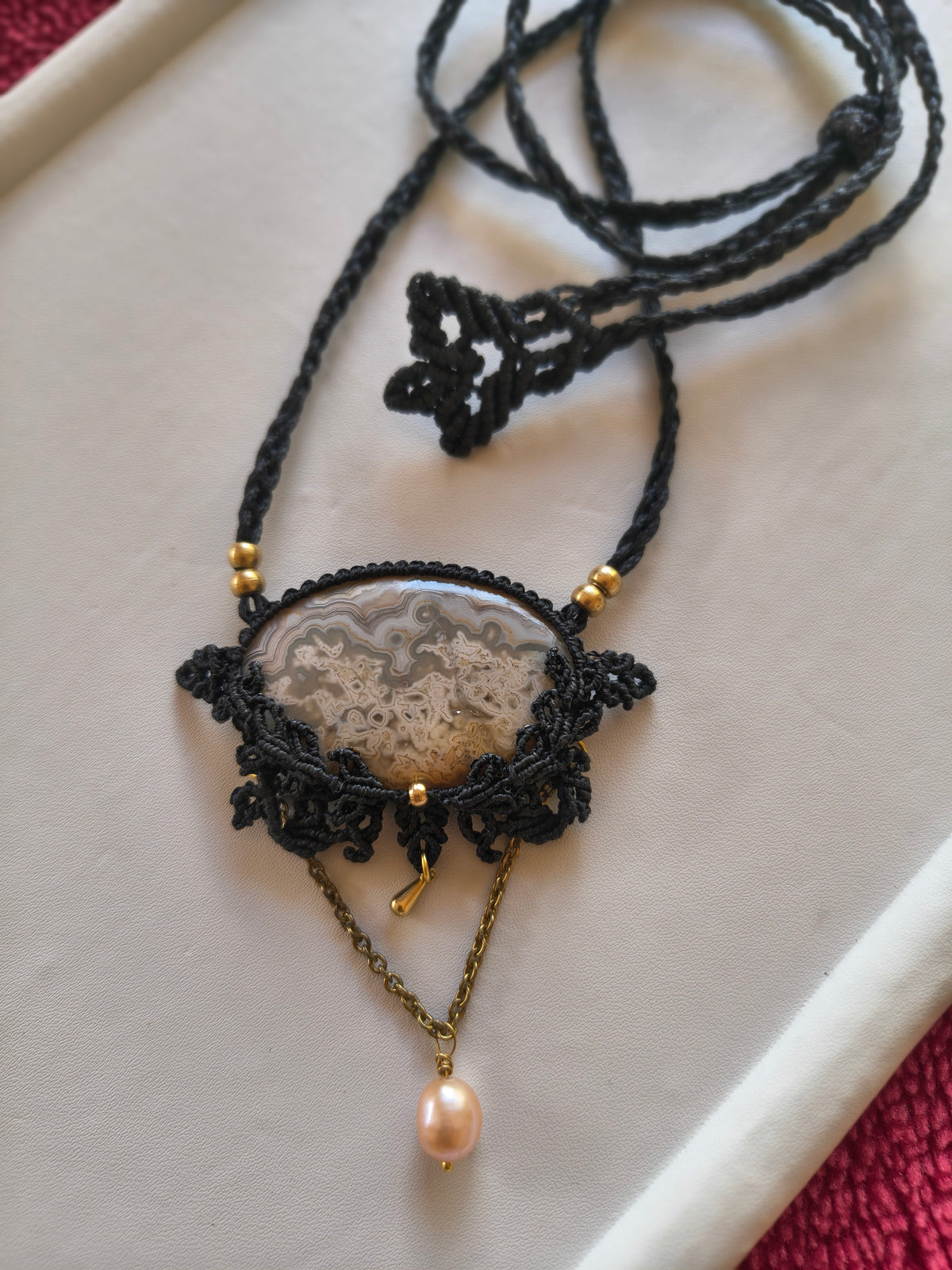 A handmade macrame pendant necklace featuring a polished agate slice, gold-tone beads, and a dangling pink freshwater pearl on a gold chain, displayed on a white tray.