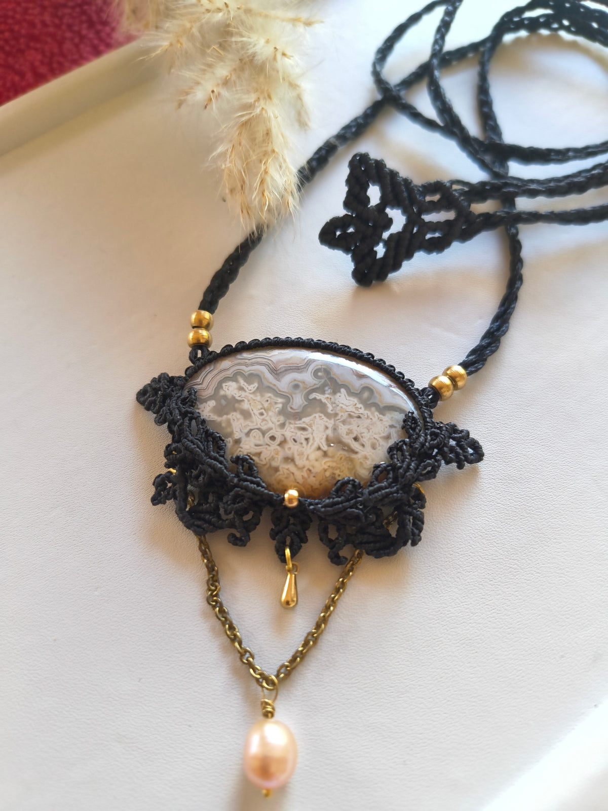 A bohemian-style macrame pendant necklace featuring a polished crazy lace agate stone in a black woven macrame, accented with gold beads and a dangling gold chain with a pink pearl, displayed on a light beige surface.