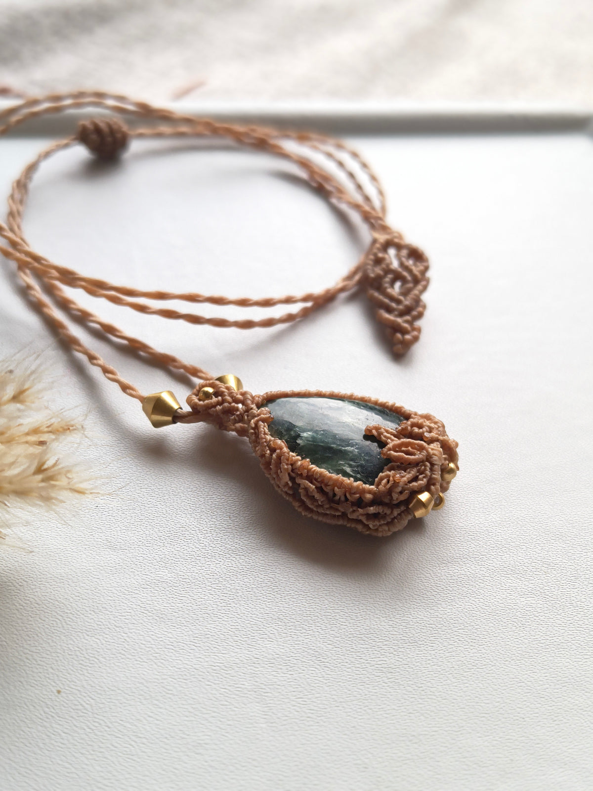 A handmade macrame pendant necklace featuring a dark green teardrop-shaped stone wrapped in brown cord, with small gold spacer beads, displayed on a light textured surface.